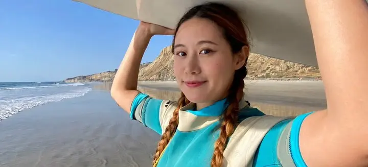 奎嵐綁著雙辮、穿藍白防寒衣,在加州海灘把衝浪板舉在頭頂,身後是海浪、沙灘與峭壁